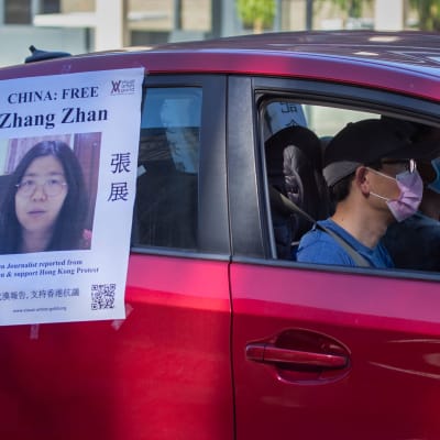 Två personer i munskydd sitter i en röd bil. På bilen hänger en lapp med en bild på journalisten Zhang Zhan och en uppmanan att fria henne från fängelset.