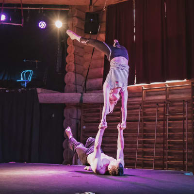 Kaksi sirkustaiteilijaa tekemässä temppua, nainen seisoo käsillä selällään maassa makaavan miehen käsien varassa