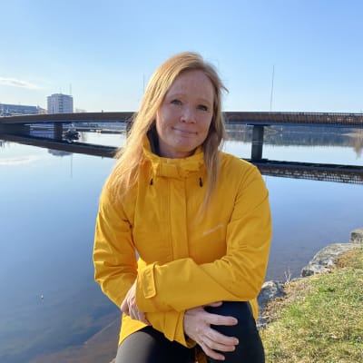 Tanja Lajunen istuu, katsoo kameraan ja hymyilee. Hänellä on yllään keltainen ulkoilutakki. Taustalla näkyy järvi, silta ja nurmikkoa.