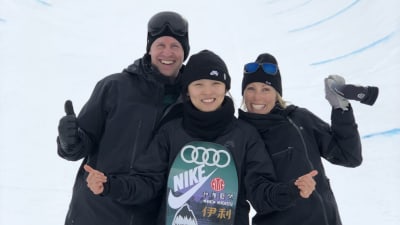 Tre personer tittar mot kameran och ler stort. Personen i mitten håller i en snowboard. De står i en snötäckt "halfpipe" som är en slags bana för snowboarding.