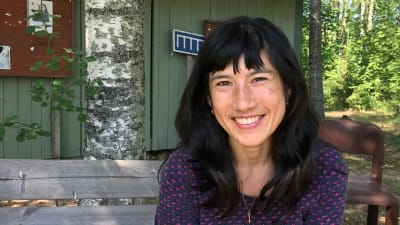En svarthårig leende kvinna med bruna ögon som sitter på en bänk utomhus.