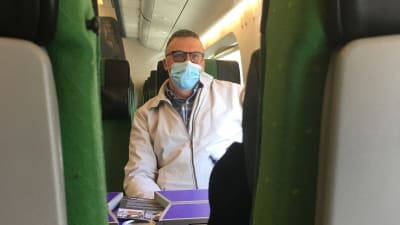 En man med kort hår, glasögon och munskydd sitter i ett tåg.
