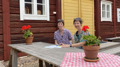 Susanne Lagus och Gunilla Sand sitter utomhus vid ett bord med papper framför sig på bordet och en röd stockvägg bakom sig.
