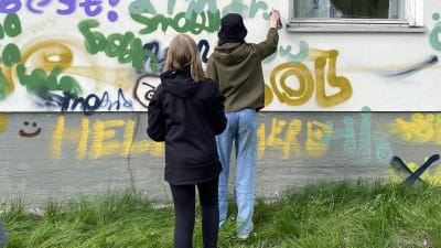 Två unga tjejer sprejar grafitti på grå vägg.