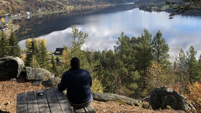En kvinna sitter på en bänk ute, ryfggen vänd mot kameran. Höst, sol och utsikten är en fjord, hus vid stranden och fjäll i bakgrunden. Norge, Hols kommun.