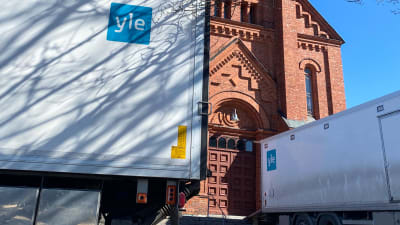 Två stora vita lastbilar med Yle-logo framför en kyrka i rött tegel.