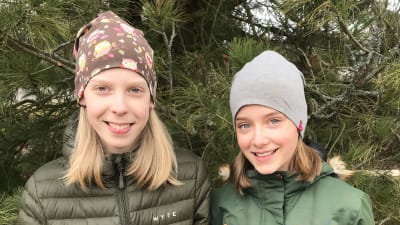 Två flickor står utomhus och ler medan de tittar in i kameran. De är klädda i jacka och mössa.