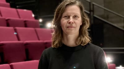 En kvinna med axellånt hår och svrat tröja framför röda bänkrader.