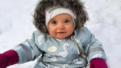 En bebis i blå vinteroverall sitter i en snödriva och ler.