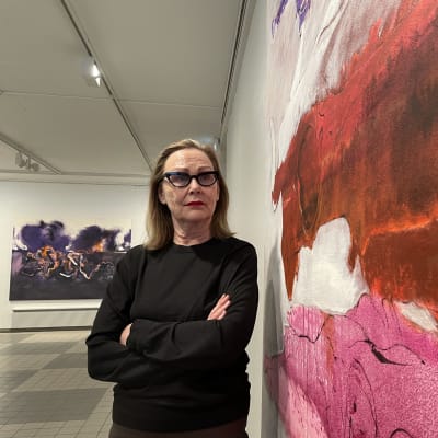Taiteilija Marjatta Tapiola seisoo suuren taidemaalauksensa vieressä kädet puuskassa taideteosta tutkien.