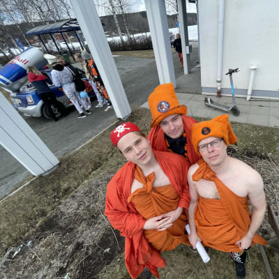 Oransseihin toogiin pukeutuneita nuoria miehiä seisoo lumesta vapautuneella, harmaalla nurmikolla. Taustalla opiskelijahaalareihin sonnustautuneita henkilöitä.