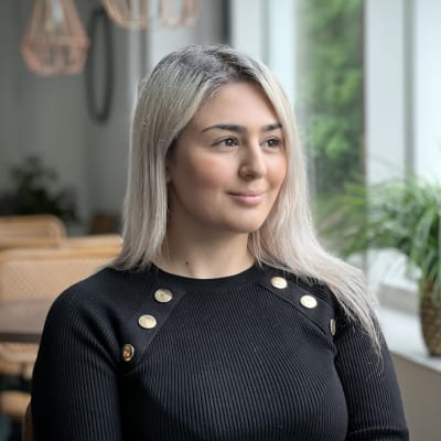 Kokemusasiantuntija Samira Nouri musta paita yllään katsoo kohti ikkunaa, taustalla ikkunalaudalla viherkasvi. 