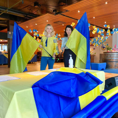 Kaksi naista pitelee isoja Ukrainan lippuja käsissään. Naisten edessä pöydällä on lisää lippuja, ja taustalla näkyy keltaisia ja sinisiä koristeluita.
