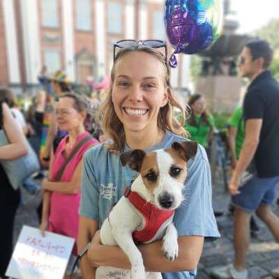 En ung dam med ljusblå skjorta , solglasögon i pannan och ett stort leende håller en brunvit liten hund i famnen.