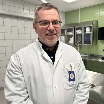 Valkoisiin pukeutunut patologian ylilääkäri Markku Aaltonen leikkasalin vihreän seinän ja teräspöytien edessä.