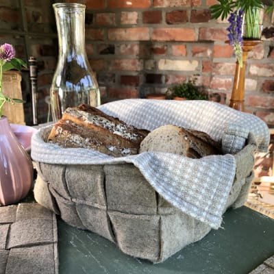 En korg gjord av flätad linfilt står på ett bord. I korgen finns en handduk samt uppskuret bröd. På bordet står även en glasflaska. I bakgrunden syns en tegelvägg.