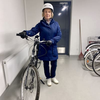 Sinitakkinen nainen, jolla on valkoinen pyöräilykypärä päässään seisoo kellaritiloissa sähköpyörän kanssa. Tilassa on valkoiset seinät ja naisen takana sininen ovi.