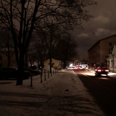 En mörk gata i Borgå centrum. Till vänster träd, till höger hus. På gatan kör en bil.