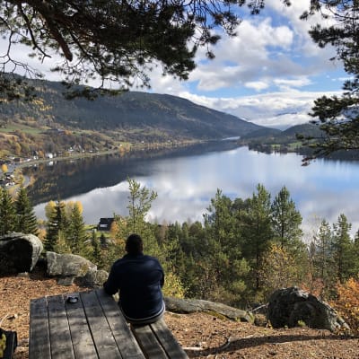 En kvinna sitter på en bänk ute, ryfggen vänd mot kameran. Höst, sol och utsikten är en fjord, hus vid stranden och fjäll i bakgrunden. Norge, Hols kommun.