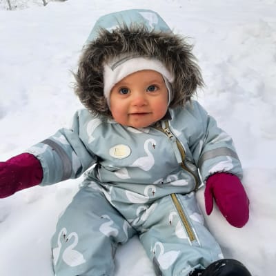 En bebis i blå vinteroverall sitter i en snödriva och ler.