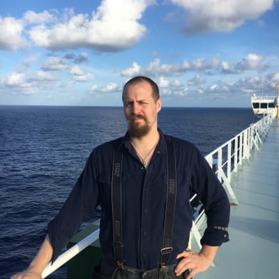 en skäggig man som står på däck på ett fartyg och i bakgrunden ser man havet.