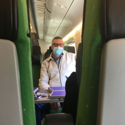 En man med kort hår, glasögon och munskydd sitter i ett tåg.