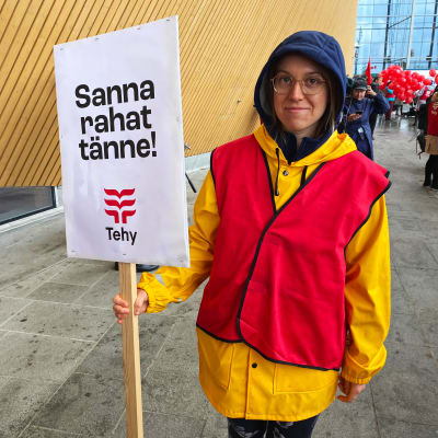 En sjukskötare i regnkläder som håller i en skylt med texten "Sanna pengarna hit!".