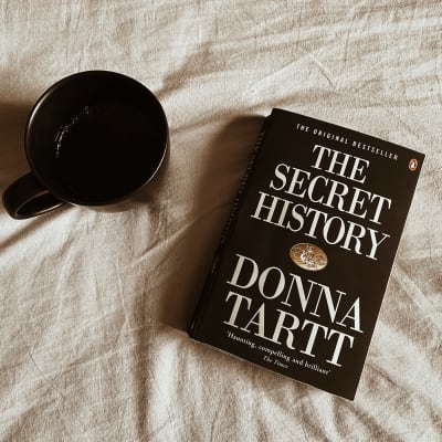 En kaffekopp och boken "Den hemliga historien" ligger utspridda på ett lakan. 