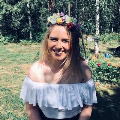 En kvinna med blont hår sitter ute i solen och skrattar. Hon har en vit blus på sig och en stor blomkrans på huvudet. Det är sommar och i bakgrunden syns björkar. 