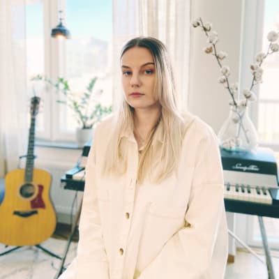 Josefin Sirén sitter ner iklädd i en vit skjorta. I bakgrunden syns en gitarr och ett piano.