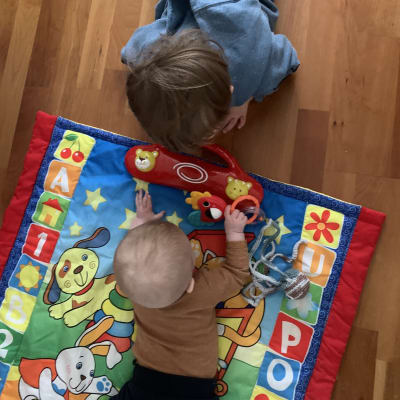 En baby och ett småbarn leker på golvet med ansiktena nedåt mot en färggrann filt.