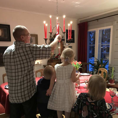 Mies sytyttää pöydän yllä olevan kynttiläkruunun kynttilöitä kolme pientä lasta vierellään penkillä.