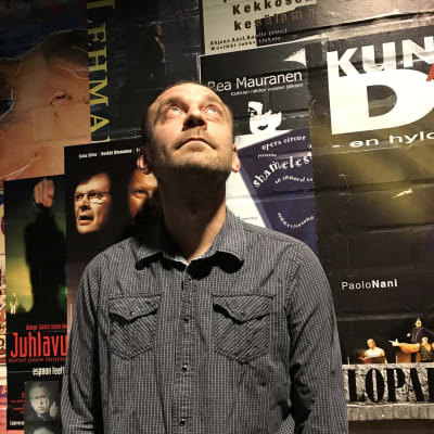 En man står framför en vägg med teaterposters och tittar upp mot en lampa. 