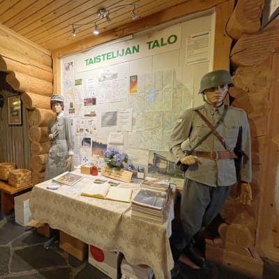 Kaksi vanhaan sotilaspukuun ja Lotta-pukuun puettua mallinukkea seisoo Taistelijan talon aulassa.