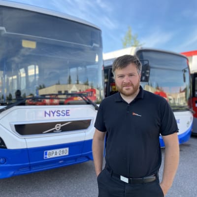 Mustaan kauluspaitaan ja tummiin housuihin pukeutunut mies seisoo sinivalkoisen Nysse-bussin edessä kädet taskussa. Aamuaurinko paistaa, taivas on sininen ja taustalla näkyy lisää busseja.