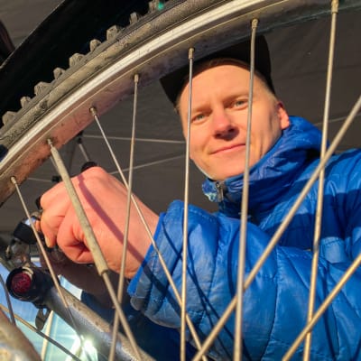 Ekokumppaneiden projektipäällikkö Pyry Rinkinen huoltaa polkupyörää ja hymyilee kameralle. Kuva on otettu renkaan pinnojen takaa.
