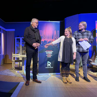 Kolme ihmistä seisoo teatterin lavalla. Mies ja nainen katsovat ihaillen vanhinta miestä, joka seisoo Paula-mainoksen vieressä.