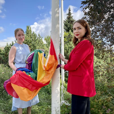 Två kvinnor i färd med att hissa en regnbågsflagga. 