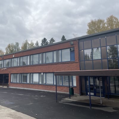 Kaksikerroksinen tiilirakennus on kuvattu sivustapäin. Koulun edessä on tyhjä piha.