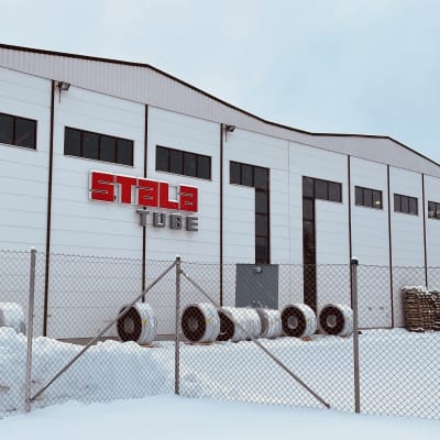 Valkoinen tehdasrakennus, jonka edustalla on lunta. Seinässä lukee punaisilla ja harmailla kirjaimilla Stalatube.