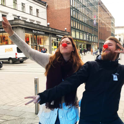 Milli Hellström och Christoffer Kaski iförda röda näsor framför en gata med människor som går.