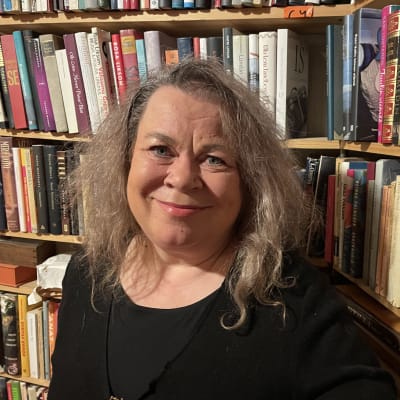 En kvinna med långt grått hår står framför en bokhylla fylld med böcker.