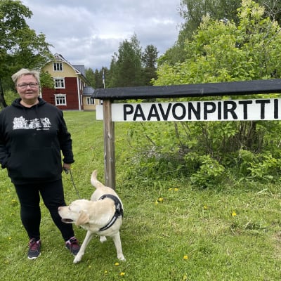 Nainen ja koira seisovat talon pihanmaalla.