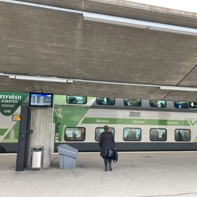 Valko-vihreä juna rautatieasemalla, nainen on nousemassa junaan.