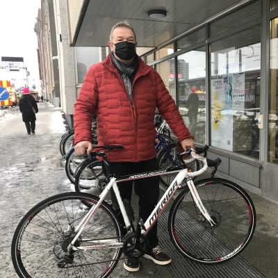 Punaiseen toppatakkiin pukeutunut Juha Suni pitelee pystyssä valkoista polkupyörää, jonka hän on tuomassa poliisin löytötavaratoimistoon. 