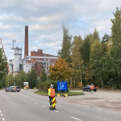 Neonvärisiin työvaatteisiin pukeutunut mies ohjaa autoja tiellä, taustalla mestää ja tehdas.
