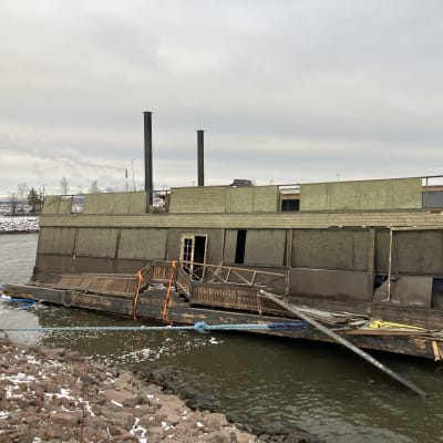 Rähjäisen näköinen puurakennelma kelluu meressä ja on kiinnitetty köysillä kiviseen rantaan.