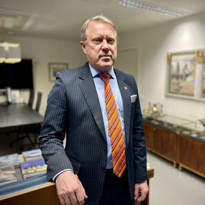 Turun yliopiston rehtori Jukka Kola nojailee työpöytäänsä tummassa liituraidallisessa puvussa ja oranssissa solmiossa.