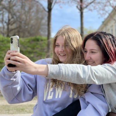 Kaksi tyttöä ottaa selfietä ja hymyilee leveästi.