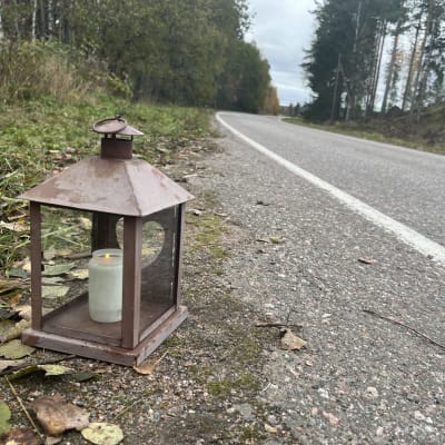 Harmaa, kaartuva asfalttitie, onka reunalla on metsää, heinittynyttä penkkaa ja keltaisia lehtiä. Tien reunassa on ruskea lyhty, jonka sisällä palaa valkoinen kynttilä.